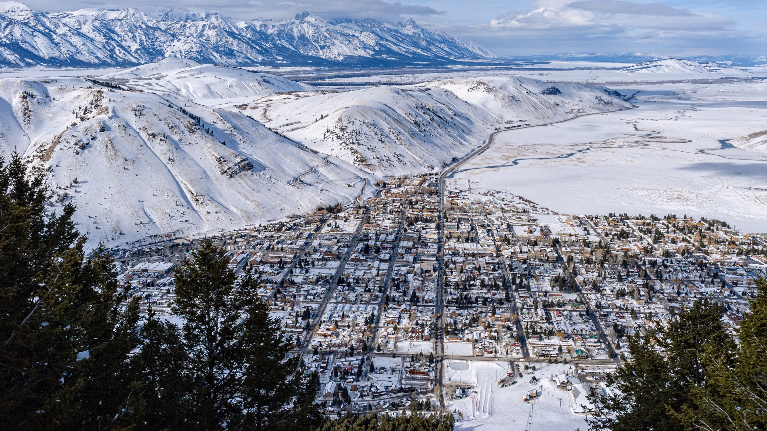 An aerial panoramic view of Jackson Hole, Wyoming, showcasing the town nestled in a valley with snow-covered mountains and ski slopes in the background