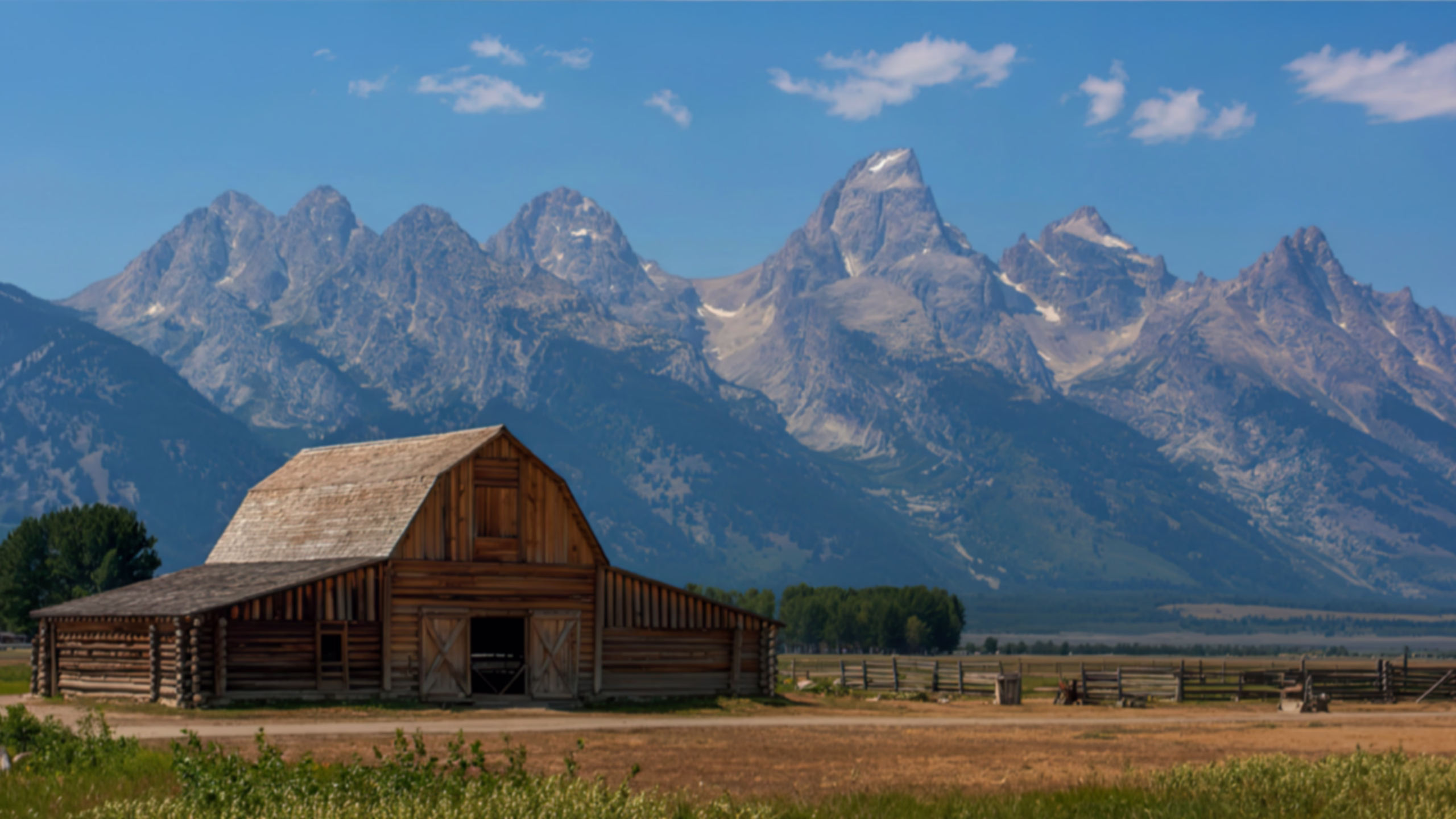 A historic wooden barn and rustic fences in a dry meadow with the majestic Teton Mountains in the background under a clear blue sky.